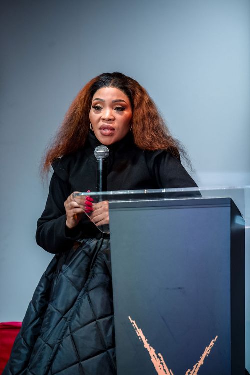 Woman with long brown hair and red nails speaking into a microphone behind a black podium.