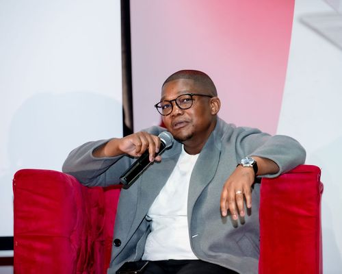 Man wearing glasses and a gray blazer sitting on a red armchair holding a microphone.