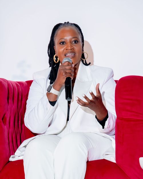 Woman in a white suit speaking into a microphone while seated on a red velvet couch.