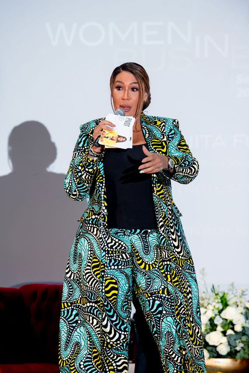 Woman in a colorful patterned suit speaking into a microphone on stage with blurred text and flowers in the background.